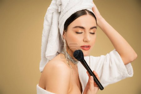 brunette woman with white towel on head applying face powder with makeup brush on beige background 