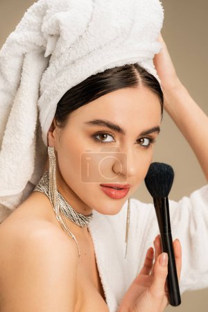 brunette femme avec serviette blanche sur la tête tenant brosse de maquillage sur fond gris 