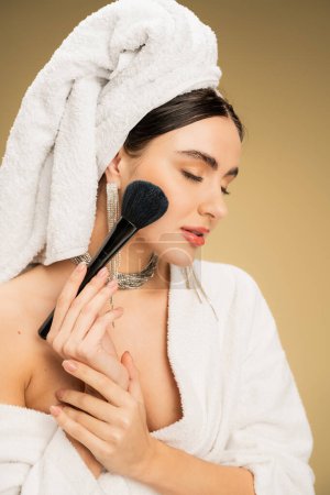 mujer joven con toalla blanca en la cabeza aplicando polvo facial con cepillo de maquillaje sobre fondo beige 