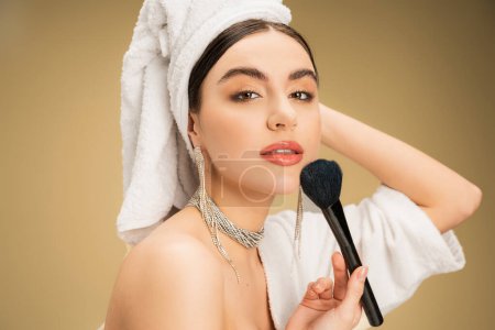 Foto de Pretty woman in white towel on head applying face powder with makeup brush on beige background - Imagen libre de derechos