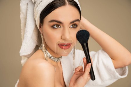 brunette woman with white towel on head applying face powder with makeup brush on grey background 