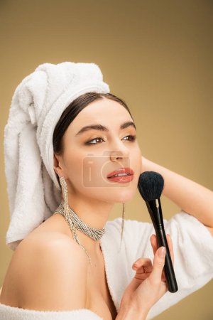 mujer encantadora en toalla blanca en la cabeza aplicando polvo facial con cepillo de maquillaje sobre fondo beige 