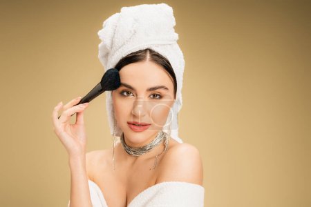 encantadora mujer con toalla blanca en la cabeza aplicando polvo facial con cepillo de maquillaje sobre fondo beige 