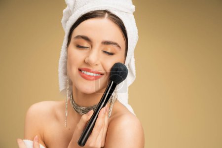 lächelnde Frau mit Handtuch auf dem Kopf, die Gesichtspuder mit Kosmetikpinsel auf beigem Hintergrund aufträgt 