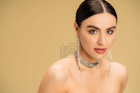 brunette young woman in necklace and earrings looking at camera isolated on beige 