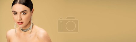 brunette jeune femme en collier et boucles d'oreilles regardant caméra isolée sur beige, bannière 