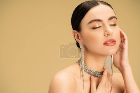 brünette junge Frau mit Halskette und Ohrringen posiert mit geschlossenen Augen isoliert auf beige 