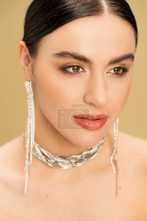 portrait of brunette young woman in necklace and earrings looking away isolated on beige 