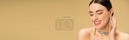 brunette young woman in necklace and earrings smiling isolated on beige, banner 