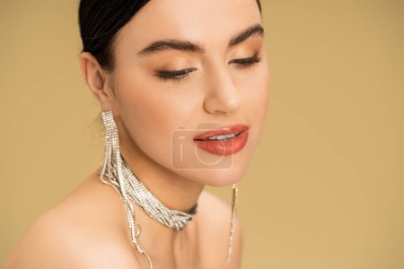 brünette Frau mit Halskette und Ohrringen schaut weg, während sie isoliert auf beige posiert 