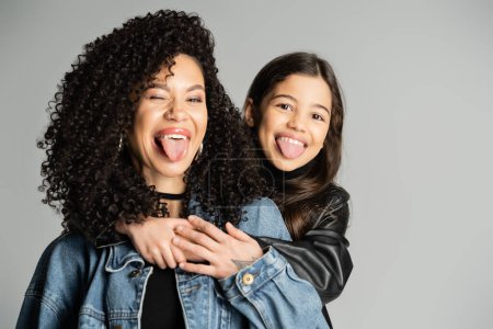Foto de Curly woman and daughter sticking out tongues isolated on grey - Imagen libre de derechos