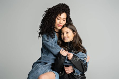 Curly woman in denim jacket hugging happy daughter isolated on grey 