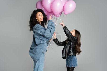 Foto de Smiling mother sticking out tongue near daughter pointing at pink balloons isolated on grey - Imagen libre de derechos