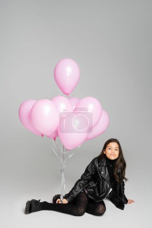 Photo for Smiling preteen kid in leather jacket looking at camera near pink balloons on grey background - Royalty Free Image