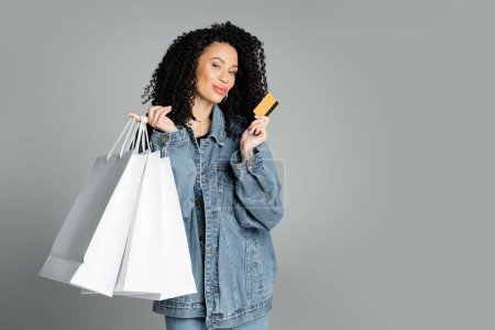 Foto de Trendy woman in denim jacket holding credit card and shopping bags isolated on grey - Imagen libre de derechos