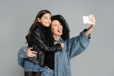 Stylish woman sticking out tongue while taking selfie with daughter isolated on grey 
