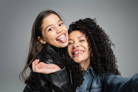 Preteen girl sticking out tongue near curly mom isolated on grey 