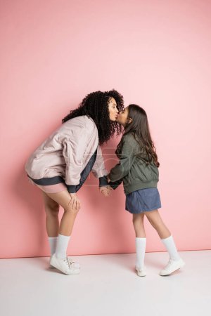 Téléchargez les photos : Side view of curly woman kissing stylish daughter on pink background - en image libre de droit