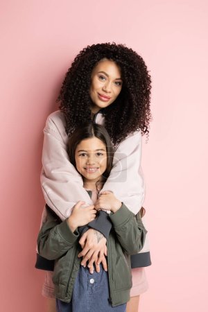 Curly woman hugging preteen girl on pink background 