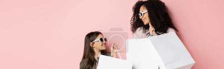 Téléchargez les photos : Cheerful woman and daughter in sunglasses holding shopping bags on pink background, banner - en image libre de droit