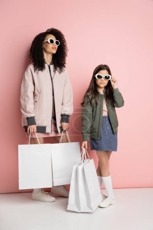Fashionable mother and kid in sunglasses holding shopping bags on pink background 
