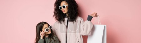 Téléchargez les photos : Trendy mother in sunglasses holding shopping bag near daughter on pink background, banner - en image libre de droit