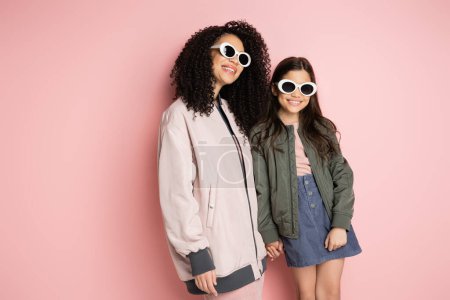 Téléchargez les photos : Positive mom and stylish daughter in sunglasses holding hands on pink background - en image libre de droit