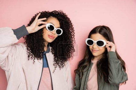 Stylish mother and daughter holding sunglasses on pink background 