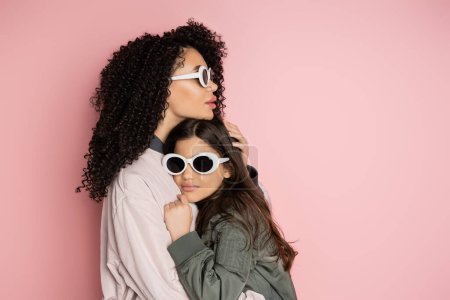 Téléchargez les photos : Stylish woman hugging daughter in sunglasses on pink background - en image libre de droit