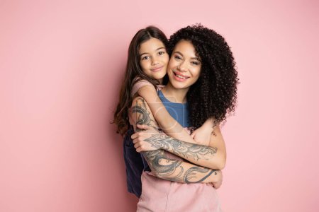 Smiling preteen girl hugging tattooed mother on pink background 