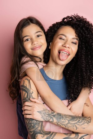 Smiling girl hugging tattooed mom sticking out tongue on pink background 