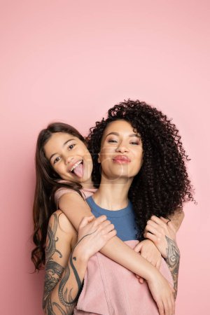 Smiling girl sticking out tongue while hugging curly mother on pink background 