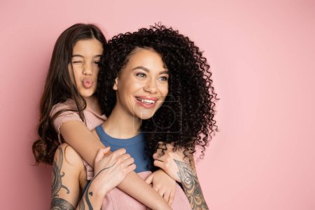 Preteen kid hugging tattooed mom and pouting lips on pink background 