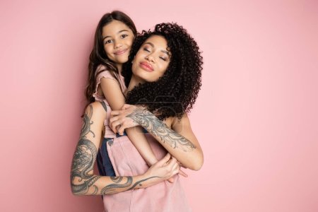 Cheerful kid hugging tattooed parent on pink background 