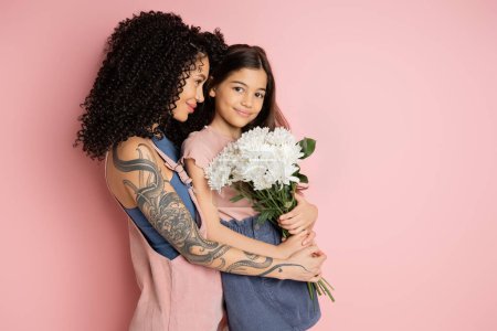 Smiling tattooed parent hugging daughter with bouquet on pink background 