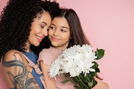 Smiling tattooed mom hugging child with white flowers isolated on pink 