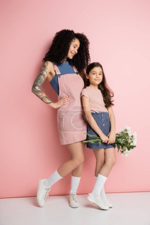 Téléchargez les photos : Pleine longueur de femme à la mode regardant fille avec bouquet sur fond rose - en image libre de droit