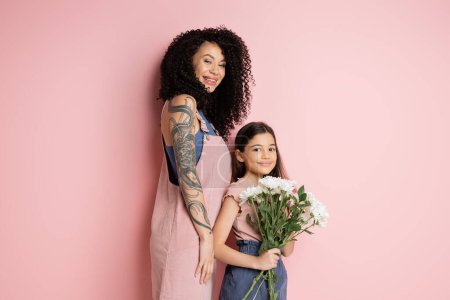 Modische Frau blickt in die Kamera nahe Tochter mit Blumen auf rosa Hintergrund 