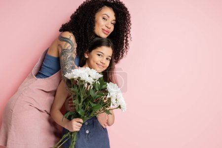 Trendy woman hugging preteen kid with flowers on pink background 