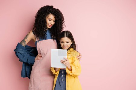 Téléchargez les photos : Trendy woman hugging daughter with digital tablet on pink background - en image libre de droit