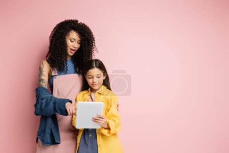 Foto de Mujer de moda apuntando a la tableta digital cerca de niño preadolescente sobre fondo rosa - Imagen libre de derechos