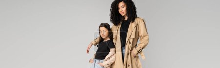 Téléchargez les photos : Stylish mom and kid in trench coats posing and looking at camera isolated on grey, banner - en image libre de droit