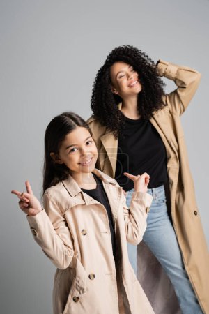 Smiling girl in trench coat showing peace sign near mom isolated on grey 