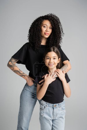 Stylish tattooed woman hugging child in t-shirt isolated on grey 