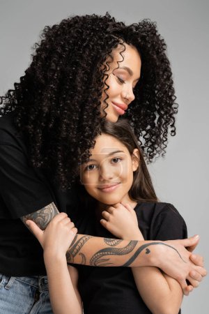 Curly woman hugging preteen daughter in t-shirt isolated on grey 