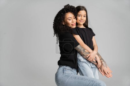 Tattooed mother hugging smiling daughter isolated on grey 