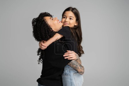Cheerful girl in jeans and t-shirt hugging tattooed mom isolated on grey 