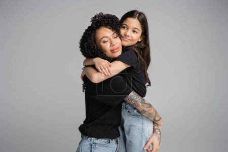 Smiling kid in t-shirt and jeans hugging curly mom isolated on grey 