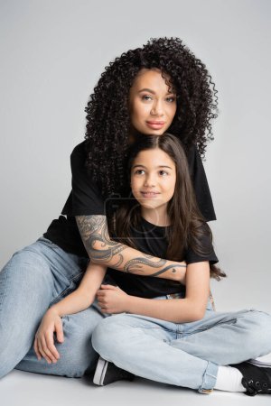 Tattooed mother hugging preteen daughter and looking at camera on grey background 