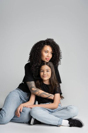 Tattooed woman hugging daughter in jeans and t-shirt while sitting on grey background 